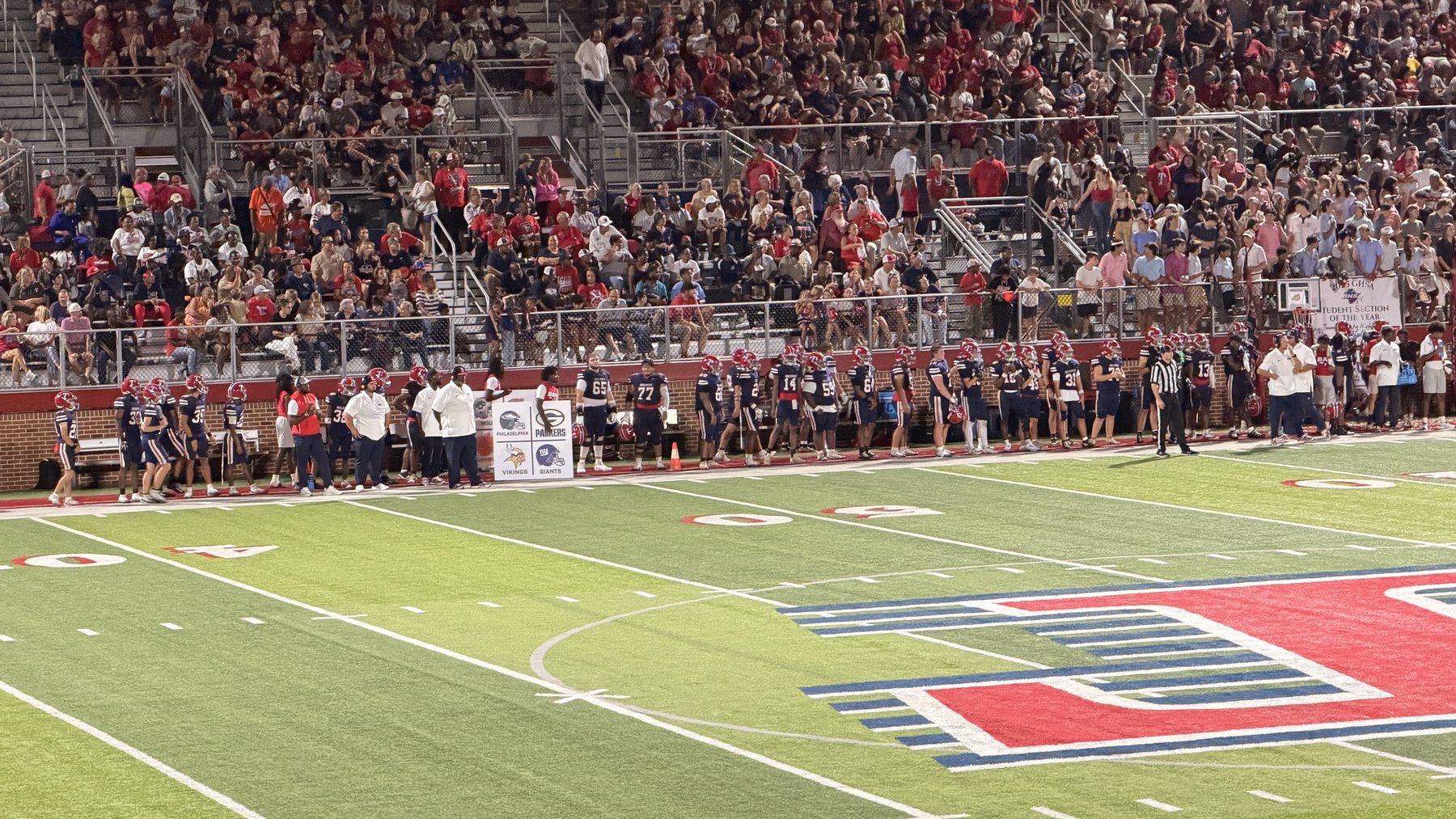 Football players along the Bulldogs sideline, with spectators filling the stands and the Flying T on the field.