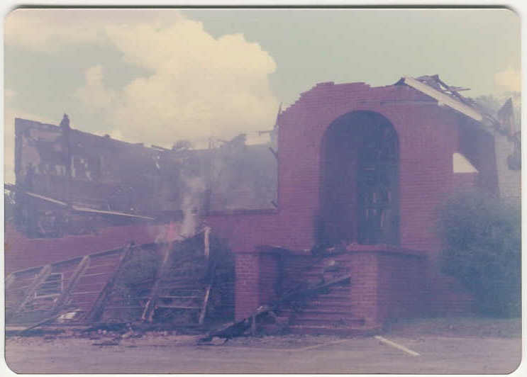 Smoke rises behind a roofless brick entrance with an arched doorway and damaged steps.