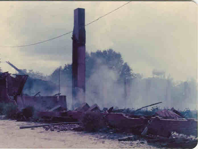 A tall chimney stands amid smoke, charred timbers, and rubble.