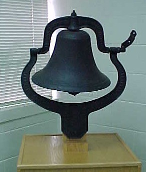 The old school bell in its metal frame, mounted on a wooden base.